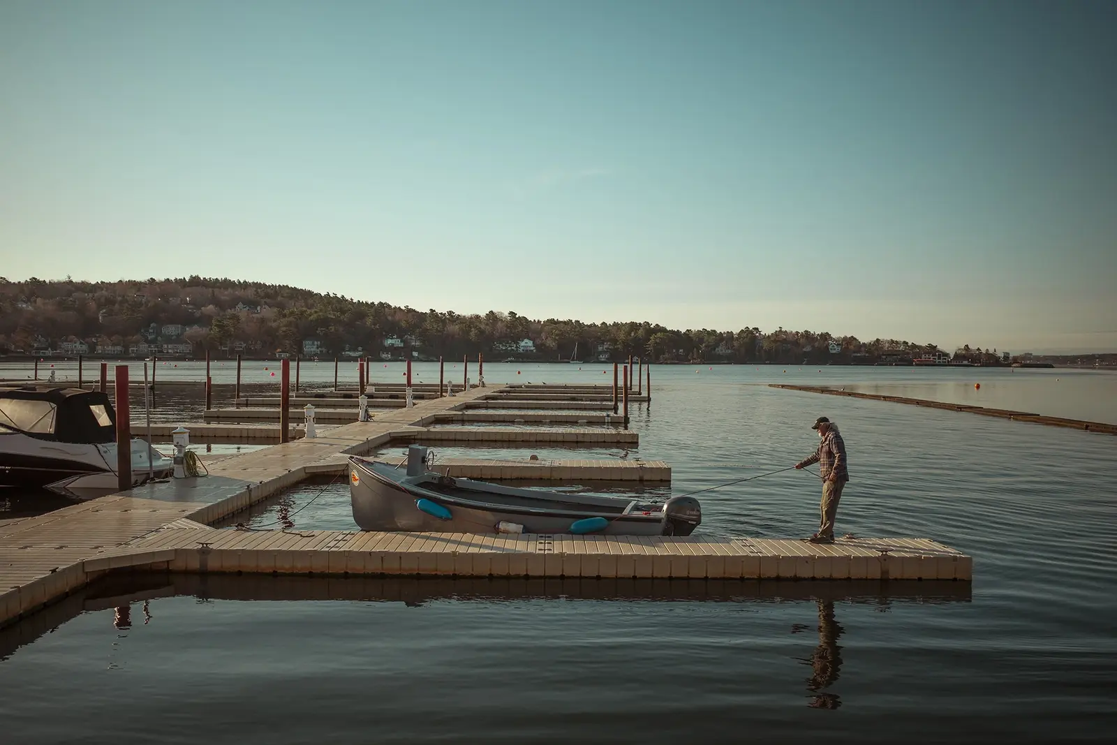 A person tending a small boat at a quiet marina, with wooded shoreline across the water